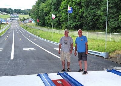 Two men stand on a soap box derby track, posing for a photo. The track is lined with flags and stretches into the distance, surrounded by trees and grassy areas. The sky is clear and sunny. Both men are casually dressed in shorts and t-shirts.